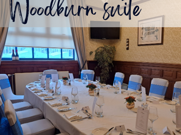 Small meeting room with large white boardroom table surrounded by white chairs with blue ribbons. Text reads Woodburn Suite.