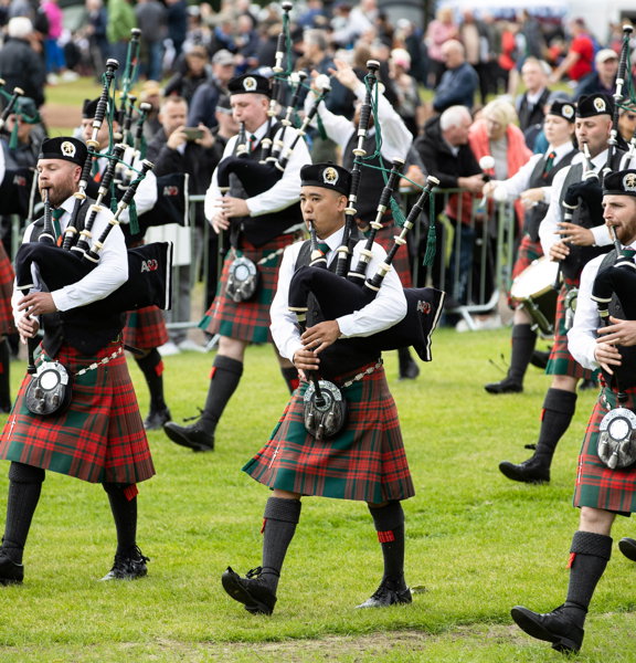 A band of pipers in red kilts march across Glasgow Green with crowds looking on