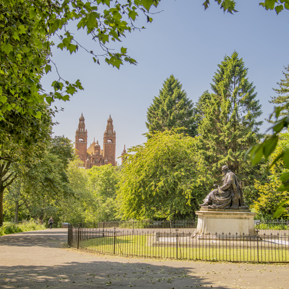 View of a large, sitting statue of Lord Kelvin, surrounded by green trees in Kelvingrove Park, with the towers of Baroque-style Kelvingrove Art Gallery and Museum in the background