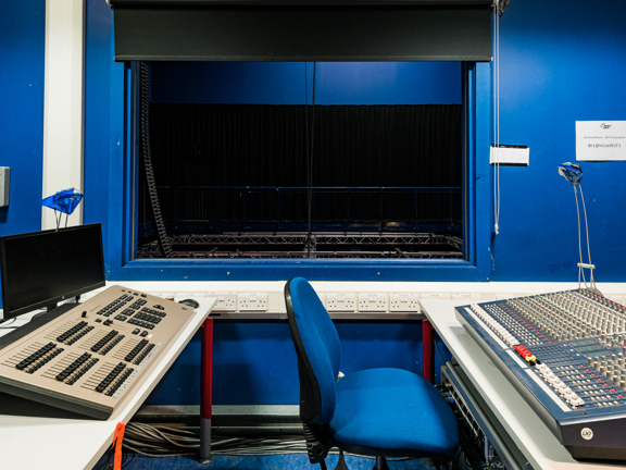 Tech studio painted dark blue with a window overlooking the theatre. Production control boards run down each side of the room. 