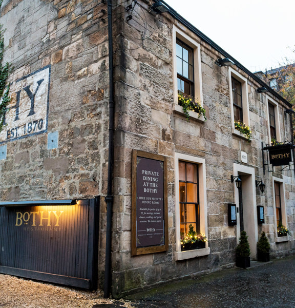 Exterior image of The Bothy restaurant shows an old, 2-storey sandstone building. The front of the building has traditional windows with dark frames, window boxes and small potted trees decorate the windows & doorway. The side of the building is half covered in ivy, obscuring a large wall painting, the "HY" of Bothy and "est.1870" are visible. A wide wooden gate, propped open against the building and a black hanging sign above the door have gold lettering reading "Bothy". The ground is paved and gravel. 