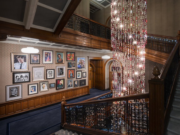 Interior view of a grand hotel staircase with dark wood panelling, ornate iron railings, a large cascading chandelier installation, and a gallery wall displaying framed portraits along a patterned wall.