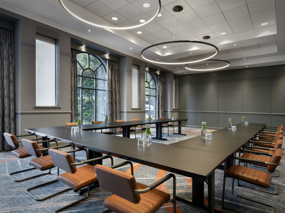 Modern and bright hotel meeting room. Wooden u-shaped table surrounded by brown leather dining chairs. Two large floor to ceiling arch shaped windows are on the left hand side of the room. 