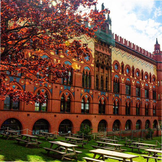 A photo of the West on the Green brewery. The facade is made of red and yellow bricks, arranged into geometric patterns. A later red brick extension with large landscape, dark-framed windows take up large portions of the visible facade. A small set of metal stairs lead down to a grassy beer garden with picnic tables.