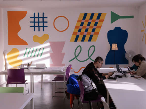 Two people sit at a white table while working on laptops. The room is brightened by a variety of colourful shapes painted on one wall. 