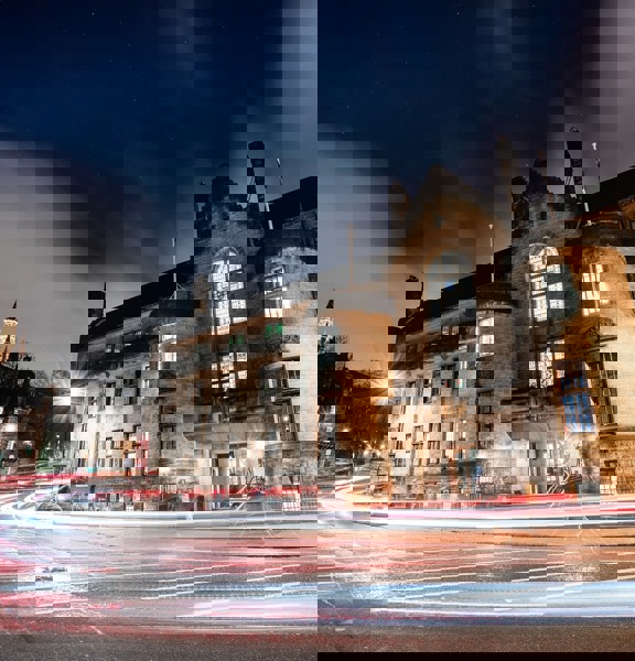 Grey stone building with turrets on either side of the wooden and glass door. Above teh door is an imposing stone balcony and large stained glass window. 