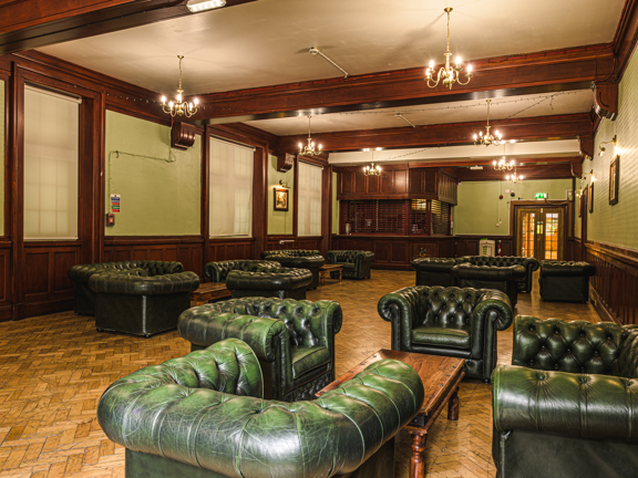 Large period room with parquet flooring, wooden panelling and beams. There are green leather chesterfield style sofas across the room around wooden coffee tables.
