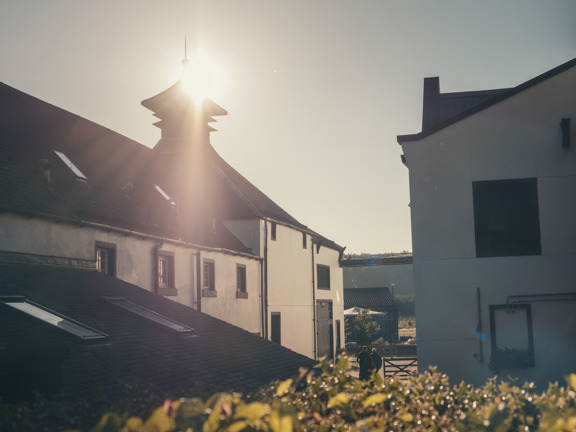 The sun is setting over a white building with a black roof, the building is part of the Glengoyne Distillery. 