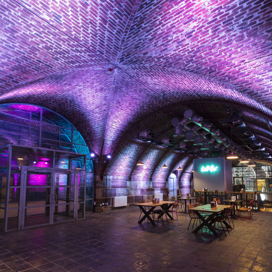 An interior image of Platform shows the junction between 2 cavernous, arched brick tunnels. The tunnel on the left has a glass and steel double doorway built into the archway., the walls on the other side are lit pink. The tunnel to the right has a tiled floor, all of the the curved walls are uplit with bright, mauve lights. Industrial-style pendant lights hang above wooden tables and wire framed chairs. A food or drink outlet at the end of the space has a turquoise neon sign on a free standing wall.