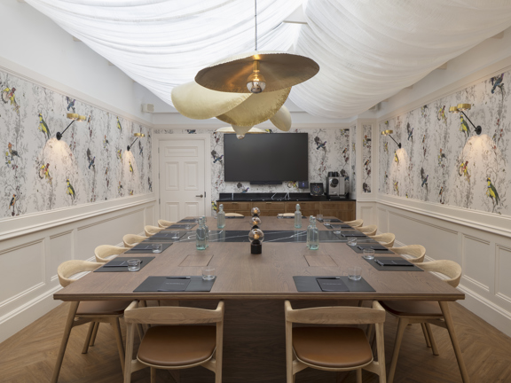 Private dining space for 22 with statement wall paper above cream wood panelled walls. Large rectangular table surrounded by wooden and leather dining chairs. Statement gold light fittings hang low above the table. 