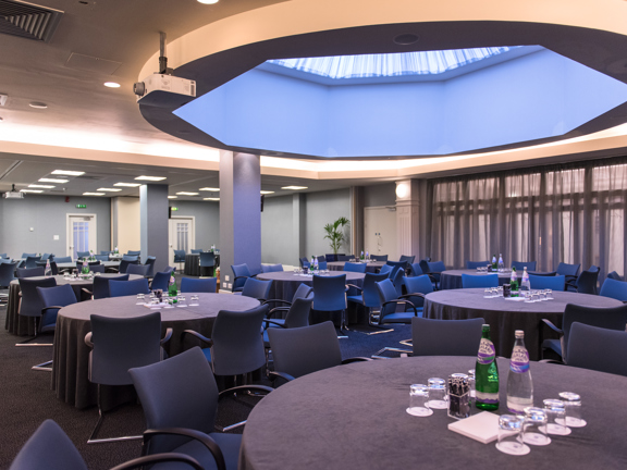 An interior image of a large room at the Golden Jubilee hotel. The room is predominantly decorated in shades of grey, and the windows on the left hand wall are covered with translucent grey curtains. Ceiling panel lights and an octagonal skylight light the room. Grey and blue fabric chairs are arranged around circular tables. They have grey table cloths, bottled water, glasses and stationary. 2 projectors can be seen in the ceiling, pointing out of shot. 3 doorways can be seen out of the room.