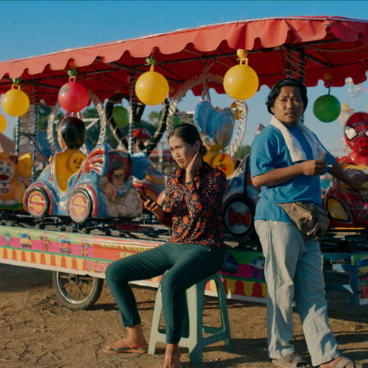 An Indonesian actor and actress sit on a colourful children's ride from a funfair.