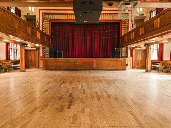 Large theatre room. The stage at front of room has red velvet curtains. There is a wooden balcony around the room at first floor level. The room has a wooden floor and natural daylight from the windows that run down either side of the room. 