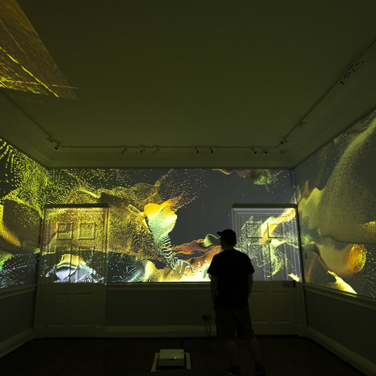 The outline of a man standing in a room with yellow, black and green pattern projected onto all walls of the room