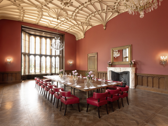 Elegant private dining room with high ornate ceiling, large bay windows with wooden blinds, and a long dining table set with glassware and flowers. The room features red walls, wood-panelled lower walls, red upholstered chairs, chandeliers, and an ornate white fireplace with a mirror above it.