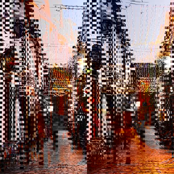 Evening view of Ashton Lane in Glasgow’s West End, with cobbled street, glowing string lights overhead, restaurant fronts and outdoor seating lining the lane, and a few blurred pedestrians walking through the scene.