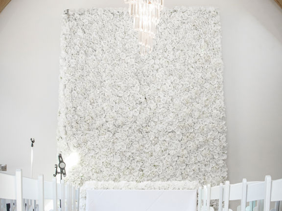 Barn function space with large chandelier hanging above rows of white chairs and a white flower wall arrangement.