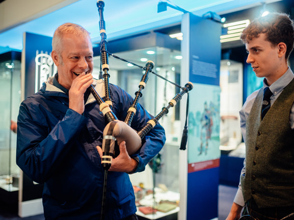A smiling man is about to blow into a bagpipe, while a smartly dressed younger man looks on