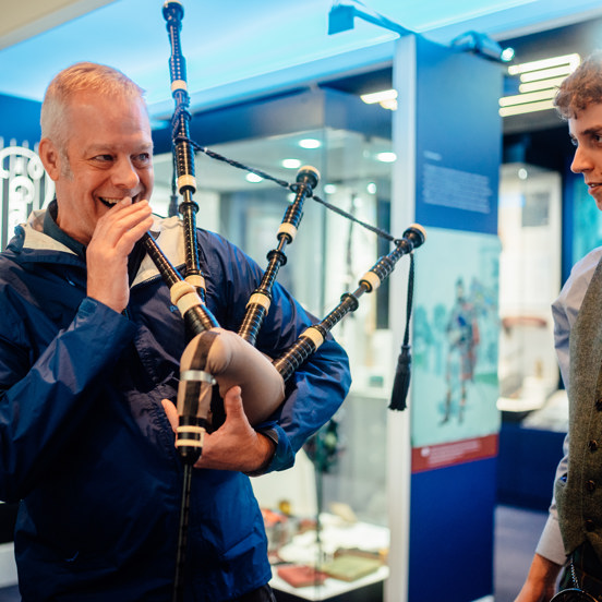 A smiling man is about to blow into a bagpipe, while a smartly dressed younger man looks on
