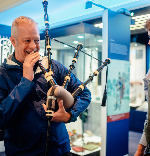 A smiling man is about to blow into a bagpipe, while a smartly dressed younger man looks on