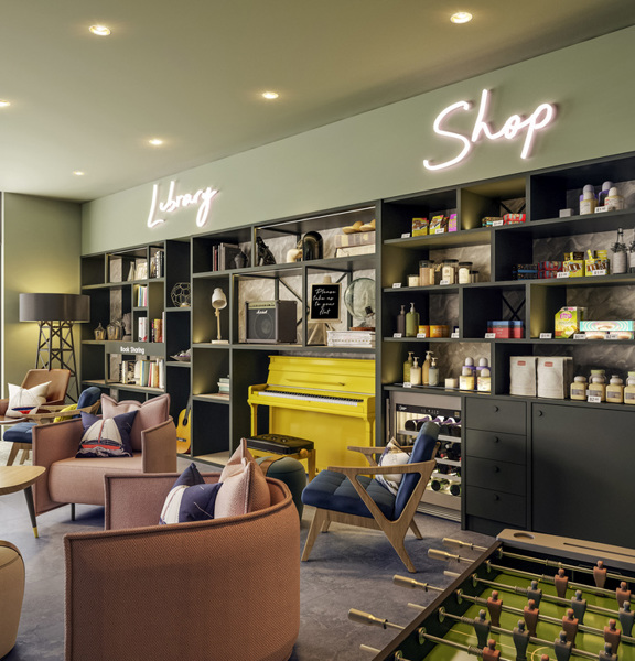 A cosy library and shop space featuring shelves filled with books and various products, including snacks and personal care items. A bright yellow piano adds a pop of colour, while comfortable seating invites relaxation and social interaction.