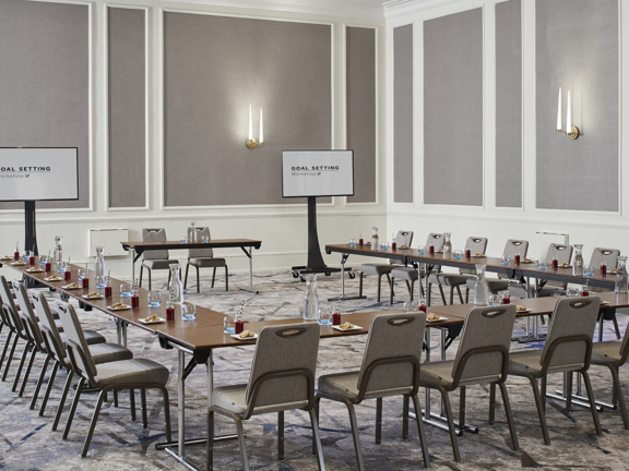 Large grey and white hotel meeting room set up with a u-shape wooden table surrounded by of grey banqueting chairs facing two large tv screens