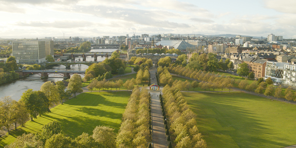 Sunny view of the tree-lined paths of Glasgow Green, on the bank of the River Clyde with 4 bridges and Glasgow city centre in the background