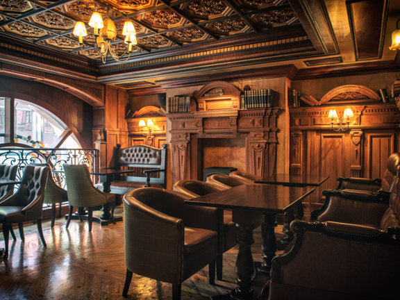 An interior image of the ornately decorated "Library" space. The room has polished wooden floors and intricately carved wall and ceiling panels. Brass ceiling lights and wall sconces with small lampshades light the space with golden light. The far, left wall is dominated by the top arch of a large window. Dark wooden tables and traditional, upholstered chairs and benches line the edge of the room, besides a large, ornate fireplace flanked by leather bound books.