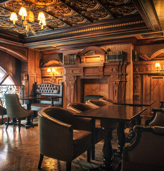 An interior image of the ornately decorated "Library" space. The room has polished wooden floors and intricately carved wall and ceiling panels. Brass ceiling lights and wall sconces with small lampshades light the space with golden light. The far, left wall is dominated by the top arch of a large window. Dark wooden tables and traditional, upholstered chairs and benches line the edge of the room, besides a large, ornate fireplace flanked by leather bound books.