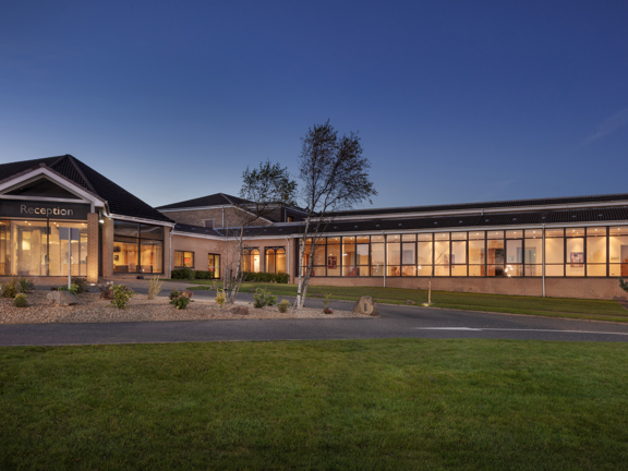 An exterior view of DoubleTree by Hilton Glasgow Westerwood, a single-storey light-coloured brick building with a dark roof. A large glass doorway with lettering reading "Reception," is on the right while a windowed-wall stretches off to the left. The sky looks like dusk so the windows glow golden in comparison. In front of the building is a paved road, a pebbled, landscaped roundabout and a lush, green lawn.