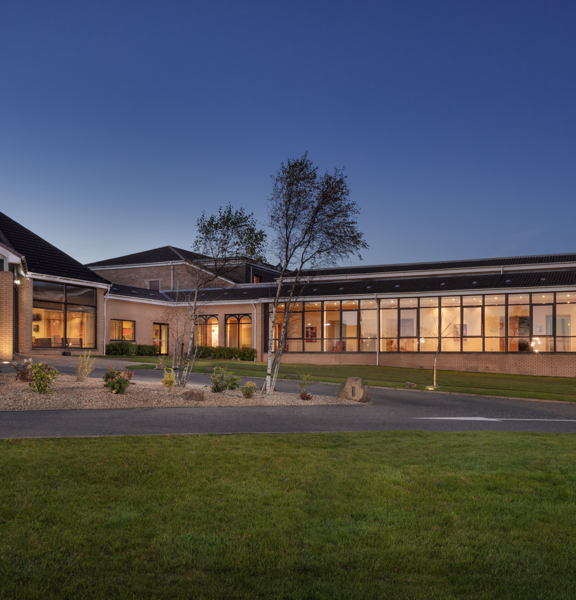 An exterior view of DoubleTree by Hilton Glasgow Westerwood, a single-storey light-coloured brick building with a dark roof. A large glass doorway with lettering reading "Reception," is on the right while a windowed-wall stretches off to the left. The sky looks like dusk so the windows glow golden in comparison. In front of the building is a paved road, a pebbled, landscaped roundabout and a lush, green lawn.