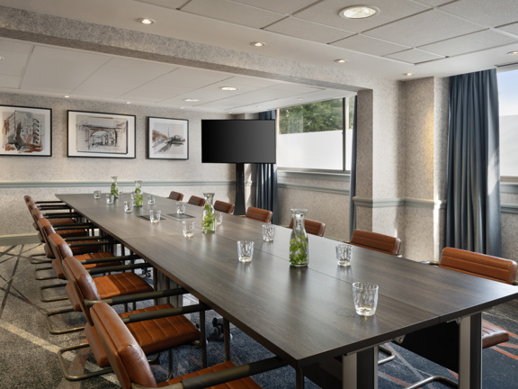 Boardroom set up in hotel meeting room. Large rectangular wooden table surrounded by brown leather dining chairs. To the right there are two large windows and a TV screen.