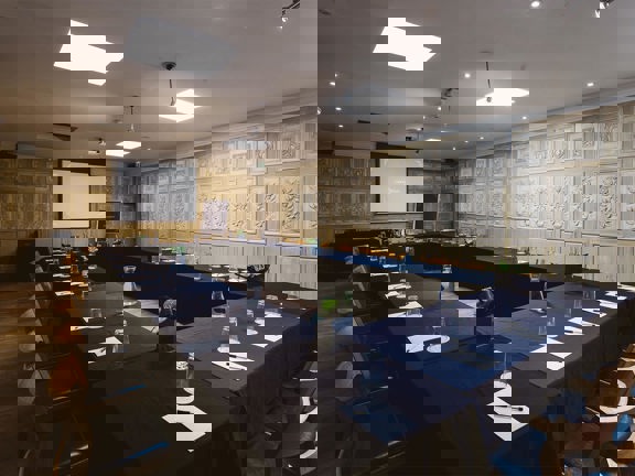 An interior view of a meeting room at Corinthian Club. 4 long rectangular tables, covered in black table cloths, are arranged into a rectangle; with wooden chairs lining the outside edge. Plants, bottles of water, paper and pens are visible on the tables. The room has a wooden floor and panelled walls. There is a large projection screen and a flipchart visible at the far end of the room. Speakers and ceiling lights are also visible.