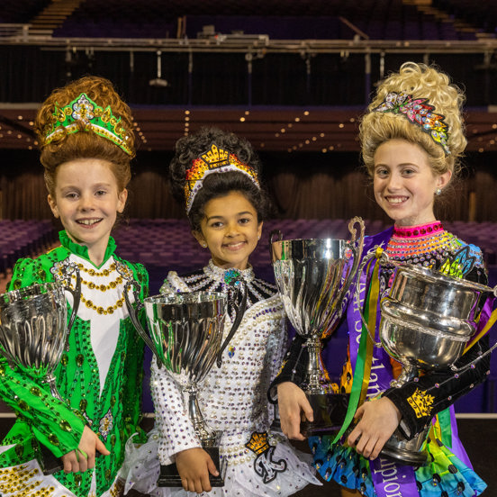 Three Irish Dancers in sparkling costumes pose with silver trophies.