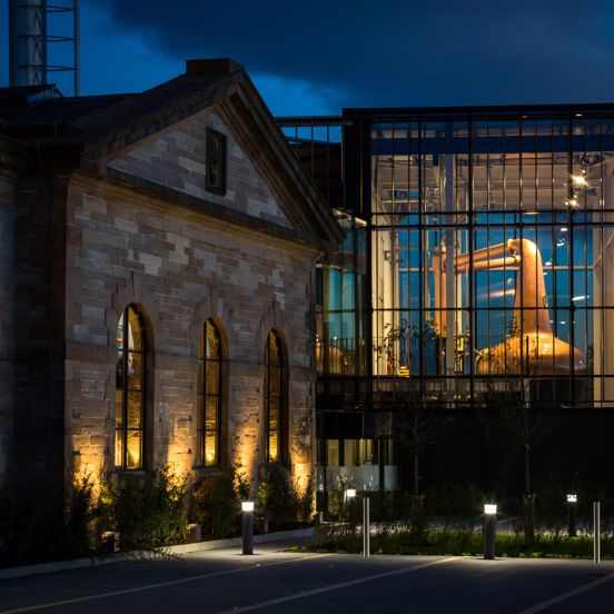 An exterior shot of the Clydeside Distillery at night, the image is taken from a tarmacked car park, some planting, outdoor lighting and silver bollards line its edge. On the left the building is old and sandstone, the base of the wall is uplit, large arched windows also glow. The right of the building is 2-storeys, modern and has glass walls. The space is lit revealing the huge, shiny copper whisky still, an enormous, bulbous instrument used in the distilling process. The sky behind is dark and cloudy.