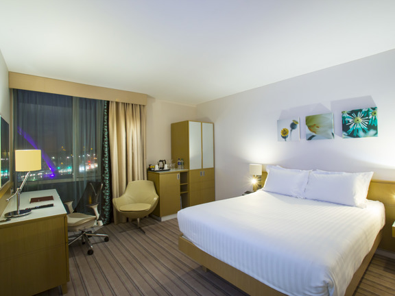 An image of a Hilton Garden Inn Queen room, taken at night, shows a room with white walls and a muted striped carpet. A bed with crisp white sheets is against the right wall, the headboard, desk, dresser and wardrobe are around the room parameters and all of the same golden wood. The large window is on the left edge of the far wall, a mesh curtain partially obscures a view of the city lights and the lit Clyde Arc bridge. A yellow leather chair, desk lamps and prints decorate the room further.