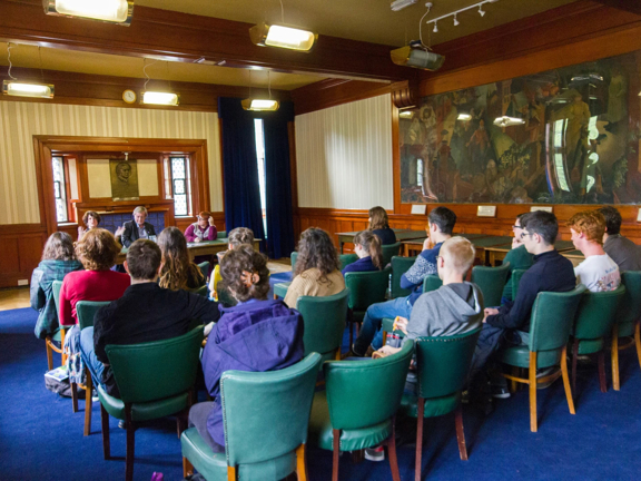 A traditional room with blue carpet and wooden panelling. People sat on rows of green leather chairs facing a panel of three people at the front of the room behind a wooden table.