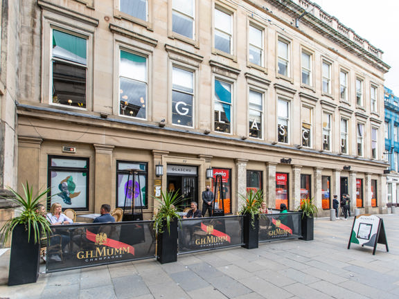 An exterior image of Glaschu's entrance. A grand, blonde sandstone building over 3 storey's, beside a paved pedestrianised area. Planters and banners separate a small private area of outdoor seating from the walkway to the left of the entrance, diners sit at 2 of the tables, either side of a stylish space heater. The first story windows of the building are decorated with colourful vinyls in organic "blobby" shapes. A white and gold letter sits prominently in each window, reading "GLASCHU".