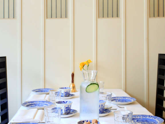 Table set for afternoon tea with blue and white linen a tablecloth and blue and white cups, saucers, plates and teapot. A backdrop of a cream wooden wall inset with stained glass windows.