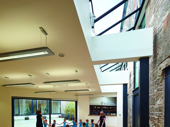 An internal view of Maryhill Burgh Hall shows a room with polished light-wood floorboards. The right-hand side of the ceiling is cut away to reveal an angled skylight.  Fluorescent lights hang from the white ceiling, supported by 2 black pillars. The right wall is exposed blonde sandstone, the far wall is diagonal and its length is a glass sliding door; a patio can be seen outside. The narrowest end of the room has a catering hatch or bar in the wall. A children's dance class is taking place in the space.