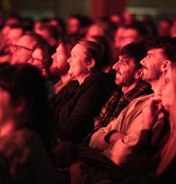 Row of men and women with red lights on their faces laughing at comedians in Barrowland Ballroom
