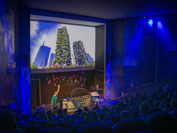 Man standing on a stage rasing his hand to a large screen displaying images of high rise buildings. Audience sits in rows of seats. 