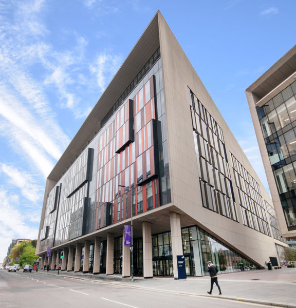 An exterior view of the TIC shows a free standing modern, angular, concrete building on a wide street. The building has a multi-storey glass facade with red and maroon geometric details. Other concrete, windowed buildings can be seen parallel and over the road. The pavement outside the building is wide and paved. A pedestrian crossing can be seen outside the building. Trees, cars, pedestrians and a bus stop can be seen farther down the road. 