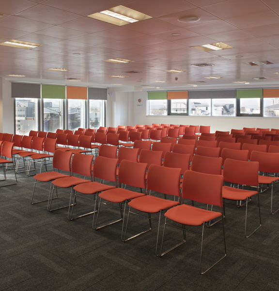 A meeting set up at The Studio shows the corner of a large room with white walls and dark grey carpet tiles. Windows with different coloured roller blinds, in orange, grey and green, wrap around the corner of the room. Dozens of red fabric chairs with wire legs are arranged into 9 rows with a central aisle. A projector is mounted on the tiled ceiling, between fluorescent lights. 