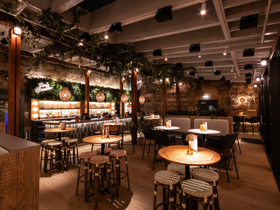 Interior of Kong bar in Glasgow featuring a cozy, modern space with wooden tables and stools, ambient lighting, hanging greenery, and a well-stocked bar against a stone wall backdrop.