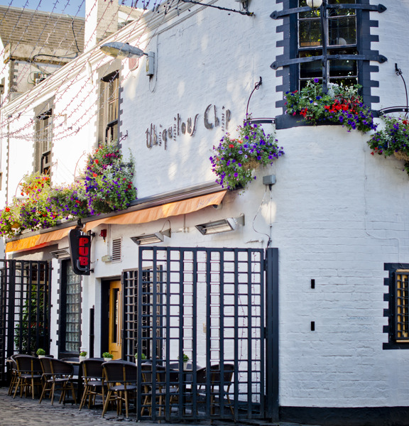 An external view of the Ubiquitous Chip restaurant's entrance off of Ashton Lane. A brick building painted white, the stone work and frames around the small, paned windows are painted black. Trellises separate an outdoor seating area, filled with small bistro tables and chairs, from the cobbled lane. Hanging baskets and window boxes bursting with colourful annuals decorate the first storey's facade. Decorative, metal lettering reads "Ubiquitous Chip" on the building's front, on the wall above the doorway.