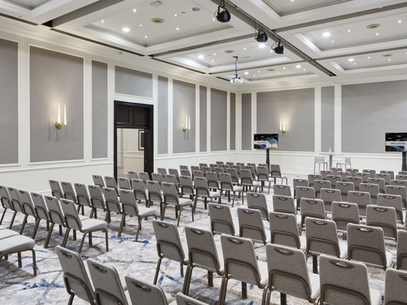Large grey and white hotel ball room set up with rows of grey banqueting chairs facing two large tv screens
