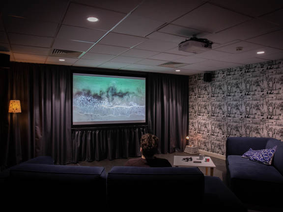 Cosy communal TV room in student accommodation. A man sits on a dark blue sofa opposite a large TV screen. The screen is mounted on a walk that is covered with a dark grey curtain. Ambient lighting gives a warm glow.  