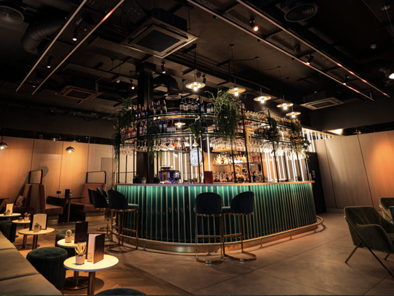 Room with a green titled bar at the centre, various tables and chairs laid out across the room. 