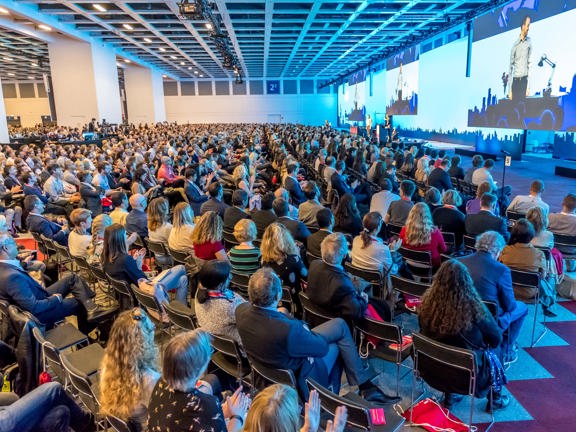Large conference room with three giant screens along right hand wall and stage set up below the middle screen.  Audience seated in rows in front of stage. 